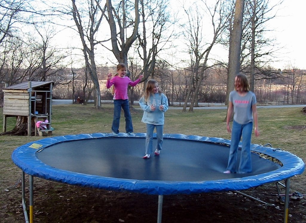 There is a group of kids. They are jumping together on a big blue rebounder. They are having a best time and they look so happy. The first girl is wearing a pink sweater, the second one is wearing blue sweater and the last one is wearing a grey t-shirt. 