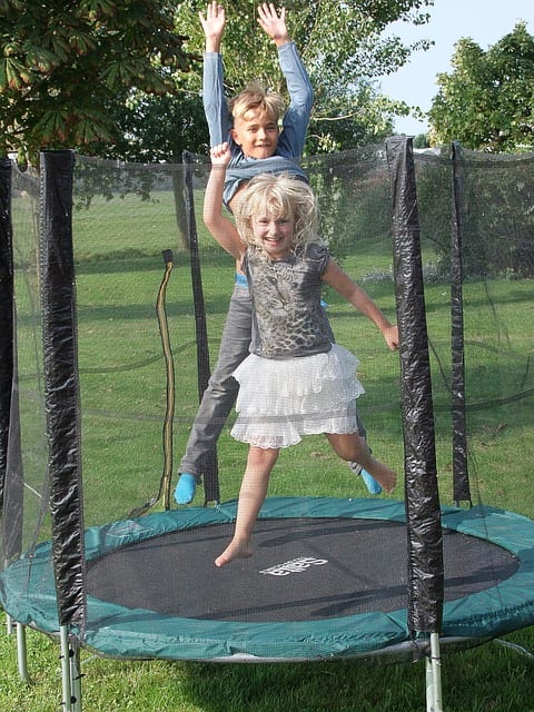 There are two children jumping together on a best rebounder. They look happy playing together. One the is wearing a pair of sock while the other is not wearing one. The rebounder has a safety net. Your children grow closer when you are happy.