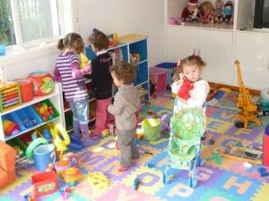 Vacuum toy: A vibrant children’s playroom filled with a variety of toys scattered across a multicolored, alphabet-themed floor mat. In the center, a child holds a toy vacuum cleaner, while other children are busy at a toy shelf and a baby doll in a green stroller, creating a lively and playful atmosphere.