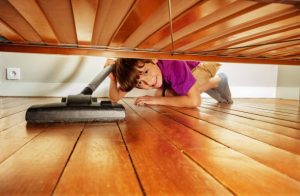 Vacuum toy: A young child lies on their stomach reaching out with a toy vacuum cleaner to clean underneath a wooden bed frame. The spacious room has polished hardwood floors, and the light filters in, casting a warm glow on the playful scene.