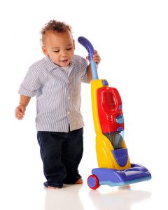 Vacuum toy: A cheerful toddler stands upright, holding onto a brightly colored toy vacuum cleaner. The toy is predominantly yellow with accents of red and blue, contrasting vividly against the plain white background. The child’s expression is one of joy and curiosity while engaging in imaginative play.