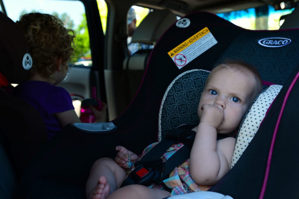 A baby sitting in a Graco car seat while sucking its thumb.