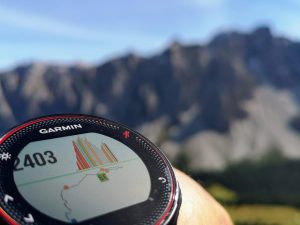 Garmin watch displaying a navigation map, worn on a wrist against a mountainous backdrop.