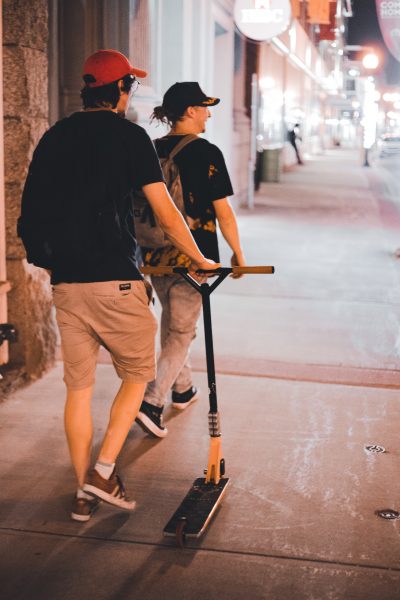 Two people in black t-shirts walk at night with a scooter without light.