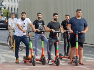 Five friends hold their scooters together while talking to each other in an urban location. 