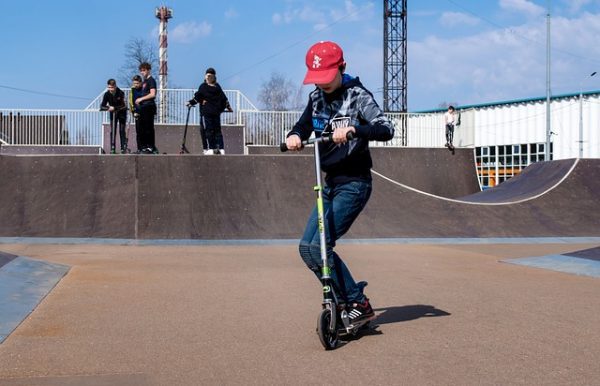 A handsome boy together with his friends at scooter park is riding his scooter and doing basic tricks at scooter ramp.