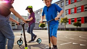 Children wearing helmets enjoying riding scooters for kids in a schoolyard. The kids' scooters are colorful. 