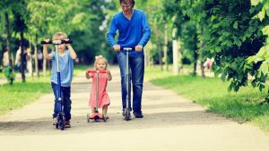 A Family enjoying a sunny day out with kids riding their own scooters in the park. 