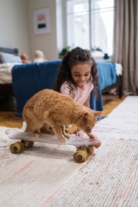 A cat riding a penny skateboard. The little girl looks very happy while watching her cat.